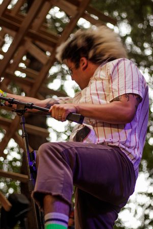 Energetic guitarist headbanging on outdoor stage, wearing striped shirt and purple pants with colorful socks.