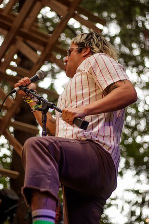 Tattooed musician with dreadlocks performs on outdoor stage, singing into microphone in striped shirt and purple pants.