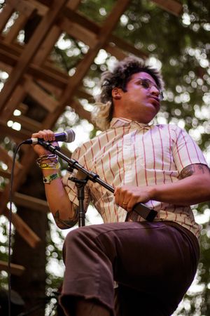 Male vocalist performing on outdoor stage, holding microphone stand, wearing striped shirt with tattoos and colorful bracelets.