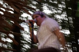 Male singer performing on stage outdoors, holding microphone, wearing striped shirt, with blurred trees in background.