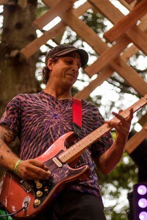 Guitarist in tie-dye shirt and cap plays red electric guitar on outdoor stage with wooden lattice backdrop.