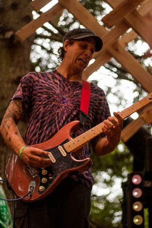 Tattooed guitarist playing red Stratocaster on outdoor stage, wearing tie-dye shirt and black cap.