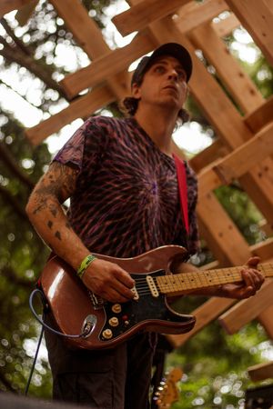 Tattooed guitarist playing electric guitar on outdoor wooden stage at music festival, wearing tie-dye shirt and black cap.