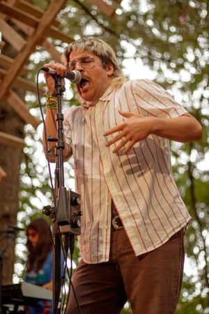 A passionate male singer performs outdoors on stage, wearing a striped shirt and glasses, singing into a microphone at a music festival.