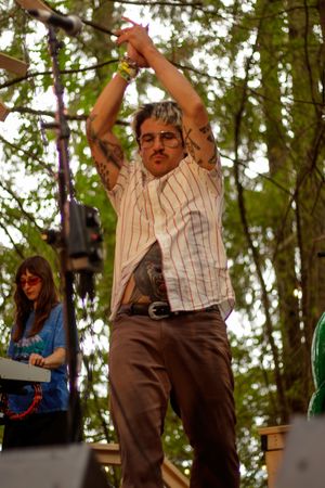 Tattooed male performer stretching arms overhead on outdoor forest stage at music festival.