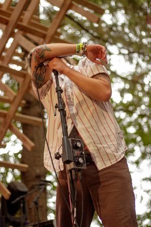 Tattooed male musician bowing head at microphone stand on outdoor wooden festival stage.