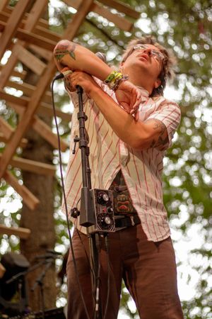 Tattooed musician grips microphone stand on outdoor stage, wearing striped shirt with vintage camera, trees visible behind.
