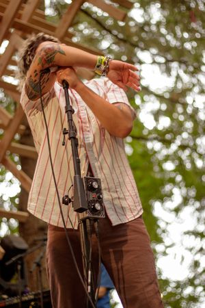Tattooed performer singing on outdoor stage, wearing striped shirt and bracelets, with microphone stand and trees in background.