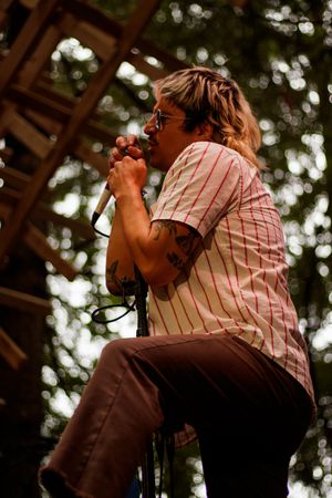 Tattooed male singer performing outdoors, holding microphone, wearing striped shirt and glasses on stage.