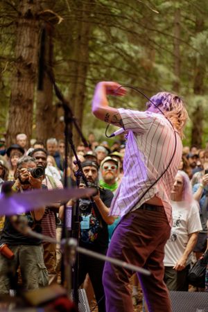 Performer with purple hair rocks out on an outdoor forest stage before a large crowd at a music festival.