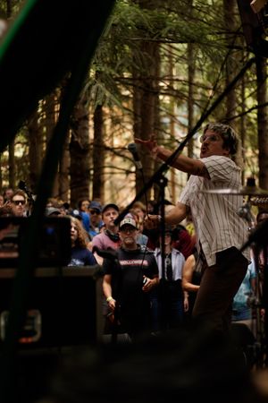 Performer on outdoor forest stage singing into microphone for crowd at woodland music festival.
