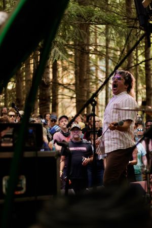 Energetic vocalist performing on outdoor forest stage to large crowd at daytime music festival.
