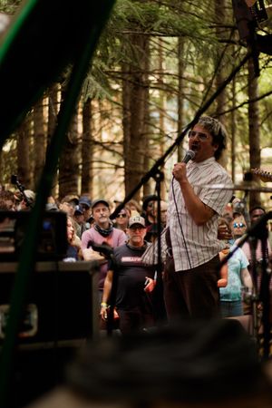 Man speaking into microphone at outdoor forest concert with crowd gathered among tall pine trees.