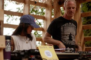 A young girl and adult man examine vinyl records at an outdoor wooden booth surrounded by trees.