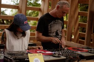 Two DJs mixing vinyl records on turntables at an outdoor music event under a wooden structure.