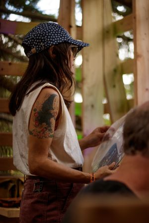 Woman with tattoos and floral cap holds vinyl record in rustic wooden structure.