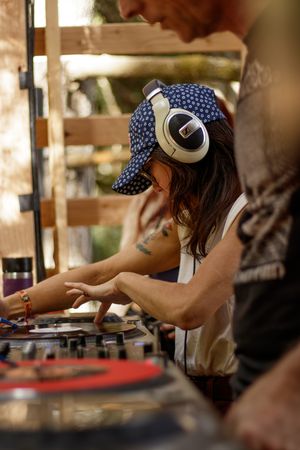Female DJ with headphones and blue cap mixing records on turntables at an outdoor music event.