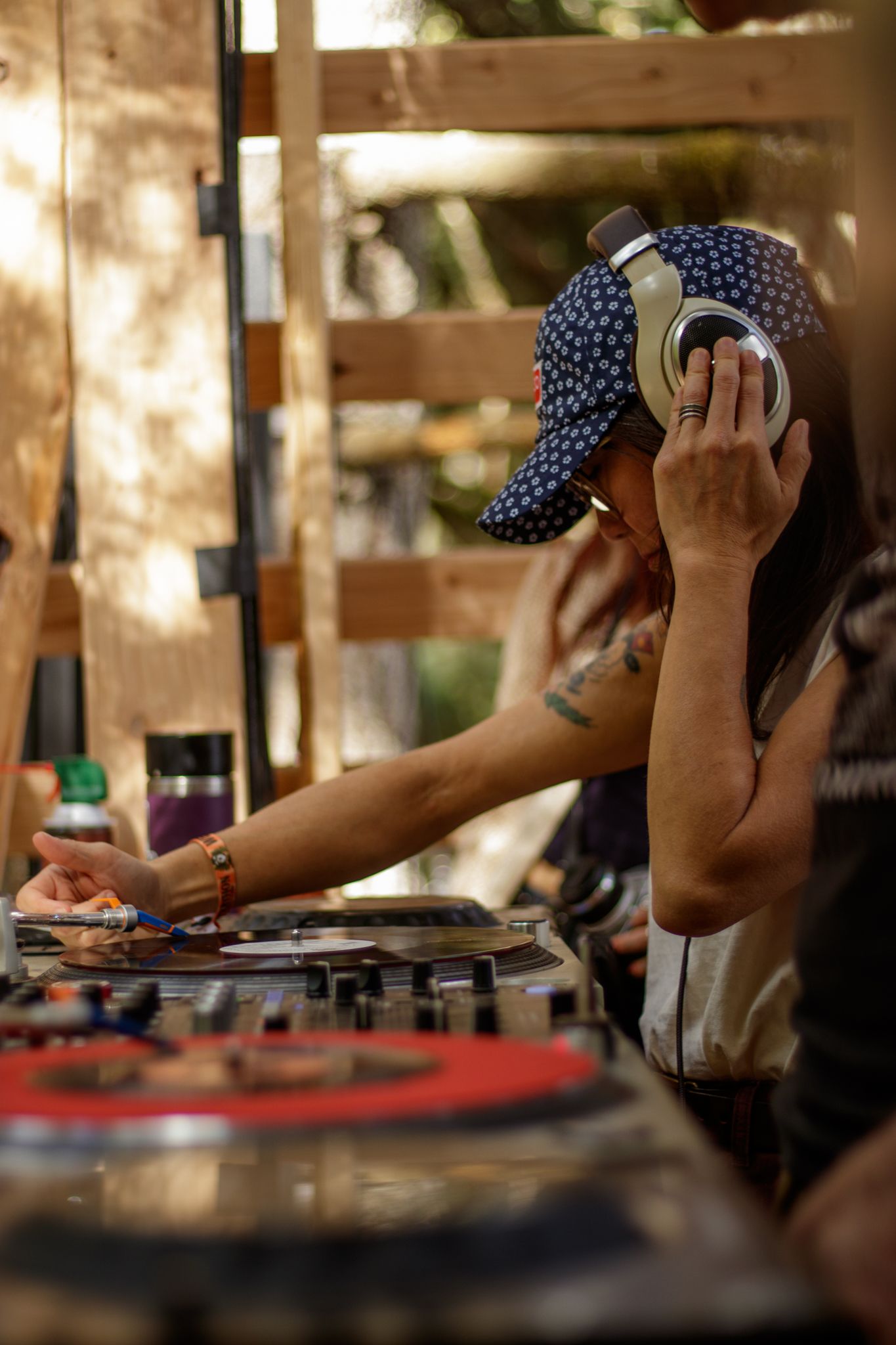 Female DJ wearing headphones and a cap mixing records on turntables at an outdoor music event.
