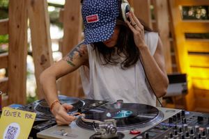 Female DJ wearing a cap and headphones mixing vinyl records on turntables at an outdoor music event.