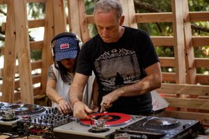 Two DJs collaborating on turntables with red vinyl records at an outdoor wooden stage venue.