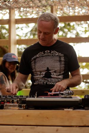 A man in a black graphic t-shirt DJs at an outdoor venue, focused on turntables under a wooden canopy.