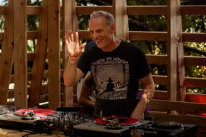 Smiling DJ waves behind turntables on an outdoor wooden stage, wearing a black graphic t-shirt.