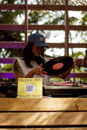 DJ handling vinyl record at Center for Cassette Studies outdoor turntable setup, wearing cap and tattoos.
