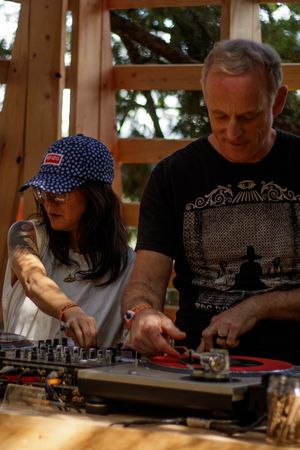 Female DJ in blue cap and male DJ collaborate on vinyl turntables at an outdoor event.