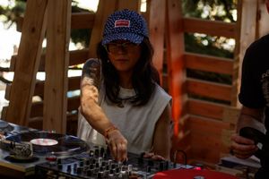 Female DJ wearing blue cap mixing records on turntables at an outdoor wooden stage event.