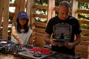 Two DJs mixing records on turntables outdoors at a wooden venue, woman in cap and man in black t-shirt.