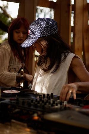 Two women DJing outdoors on turntables, one wearing a polka dot cap, in a warm sunlit wooden setting.