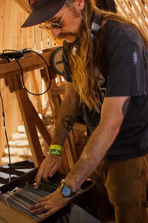 Long-haired man in black t-shirt and cap browsing vinyl records at an outdoor music festival booth.