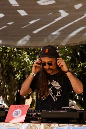 DJ LoFi performing outdoors under a canopy, wearing a Detroit Tigers cap, sunglasses, and headphones at turntables.