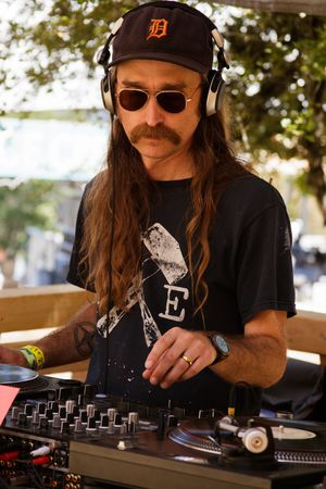 A DJ with long hair, mustache, sunglasses, and headphones mixing on turntables outdoors in a black graphic tee.