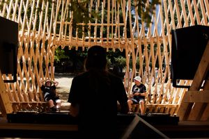 DJ performing under a latticed wooden pavilion structure outdoors with audience seated in background.