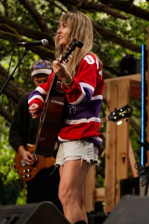 Woman in red hockey jersey plays acoustic guitar on outdoor stage with band member behind her.