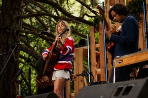 Two musicians perform outdoors on a wooden stage surrounded by trees, with a woman singing and playing guitar.