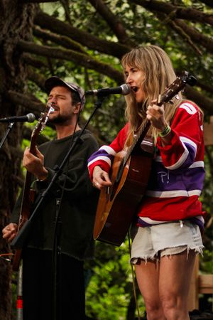 Two musicians performing outdoors, woman in red jersey playing acoustic guitar, man in black cap singing at microphone.