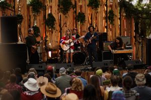 Live band performs on rustic wooden stage decorated with greenery before a large outdoor festival crowd.