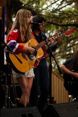 Young woman in red jersey playing acoustic guitar on outdoor stage with band members behind her.