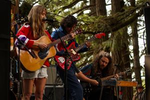 Three musicians perform outdoors on stage, playing acoustic and electric guitars among tall trees at a music festival.