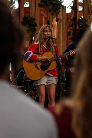 Woman in red jersey plays acoustic guitar and sings into microphone on stage before a live audience.