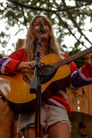 Smiling blonde woman in colorful jacket playing acoustic guitar at outdoor microphone stand.
