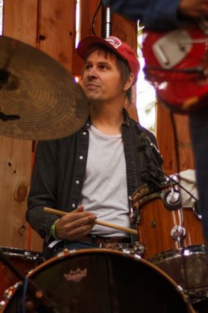 Focused male drummer holding sticks at drum set, wearing red cap, performing live on rustic wooden stage.