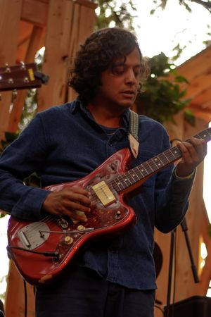 Young man with curly hair playing a red electric guitar on an outdoor stage.