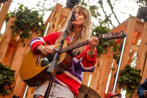 Woman in red hockey jersey plays acoustic guitar and sings at outdoor music festival.