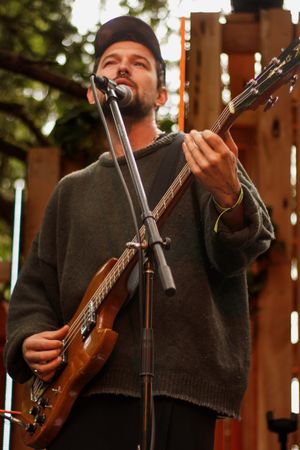 Man in gray sweater and black cap plays bass guitar at outdoor music festival.