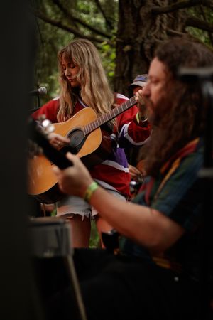 Female guitarist in red jacket performs outdoors alongside long-haired male musician at festival.