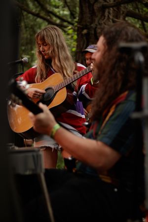Woman with long blonde hair plays acoustic guitar outdoors, with long-haired guitarist in foreground at forest music gathering.