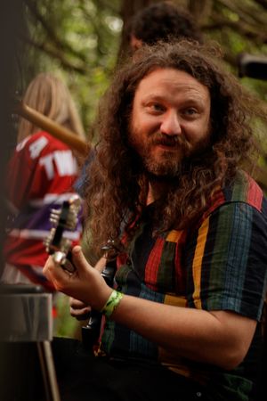 Curly-haired bearded musician playing guitar outdoors at a festival, wearing a colorful plaid shirt with a green wristband.